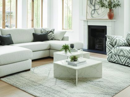 A bright, modern living room features a light grey C‑shaped corner sofa and a matching patterned armchair arranged on a neutral rug around a low marble coffee table, with large wooden‑framed windows and a minimalist fireplace creating an airy, elegant setting.