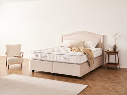 An uncovered mattress sitting on a natural coloured fabric divan set with matching contoured headboard, complete with pillows and a throw, positioned against a white wall with a white accent chair and bedside table next to it.