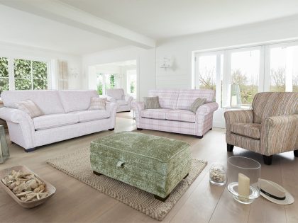 A spacious, airy living room contains a coordinated furniture set including a sofa, loveseat, armchair, and green ottoman, arranged on a neutral rug in front of large windows and glazed doors.