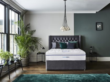 A mattress on a grey divan base with matching buttoned design headboard in a bedroom setting, positioned by a large window with plants.