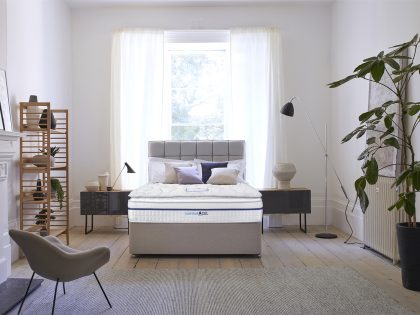 Mattress on a grey divan base with matching square designed headboard backed on to a tall window, with bedside tables either side.