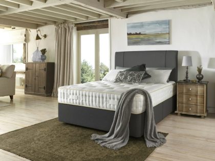 An uncovered mattress positioned on a dark grey divan base with matching headboard, sitting on a light wooden floor which has a dark green rug at the foot end of the bed.