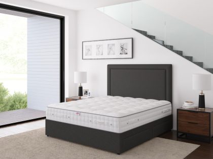 An uncovered mattress on a dark grey fabric divan base with matching headboard, placed against the wall of a staircase which can be seen through the glass balustrade, and natural light provided by the large floor to ceiling window.