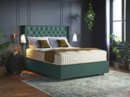 A mattress being displayed on a green divan bed with matching buttoned headboard in front of a grey panelled wall of a bedroom which features a dark wooden floor and rug.
