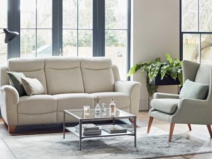 A 3 seater sofa in a light coloured fabric in front of a large window, accompanied by an accent chair and rug with coffee table sitting on it.