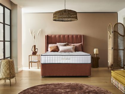 A view of a mattress and divan base from the footend, which has a matching vertical line headboard, positioned against the wall of a bedroom decorated with wicker items.
