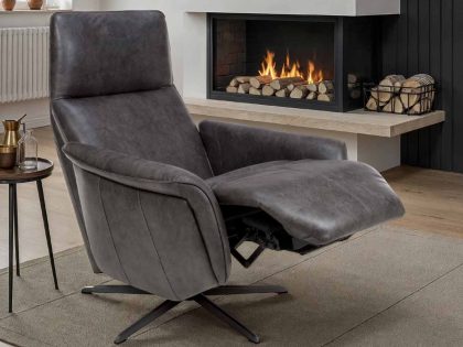 A dark grey recliner chair with a high back seen in a slightly reclined position, sitting on a rug which covers a wooden floor leading to a modern fireplace containing a lit fire.