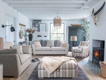 A bright, Scandinavian‑style living room features neutral sofas and chairs arranged around a patterned rug, with natural wood floors, a wood‑burning stove, and soft decorative lighting.