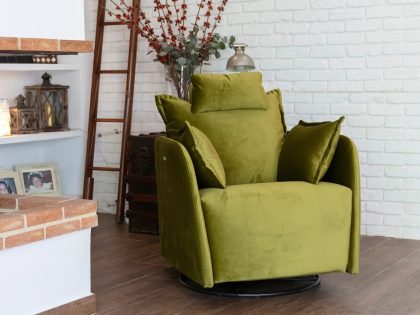 A moss‑green upholstered swivel recliner with supportive cushions is styled in a cozy living space beside a fireplace, rustic shelves, and decorative branches against a white brick wall.