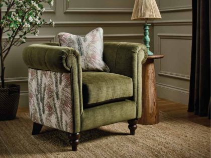 A classic accent chair upholstered in olive green fabric with patterned side panels is styled with a feather‑print cushion beside a rustic wooden side table and lamp, set against paneled walls on a woven rug for a warm, traditional look.