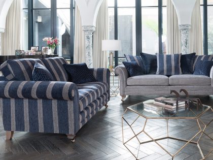 Two upholstered sofas, one striped in navy and grey and the other in soft grey velvet, face each other in an elegant, light‑filled room with arched columns, tall windows, and a geometric glass coffee table.