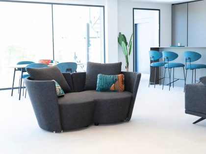 A bright, modern open‑plan living space features a curved dark grey sofa with patterned cushions, a dining table with blue chairs near large windows, and a kitchen island with matching blue bar stools in the background.