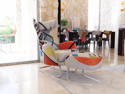 A modern living room features a brightly patterned lounge chair with a matching footstool on a rug, positioned near a glass door and a dining table with colourful chairs in the background.