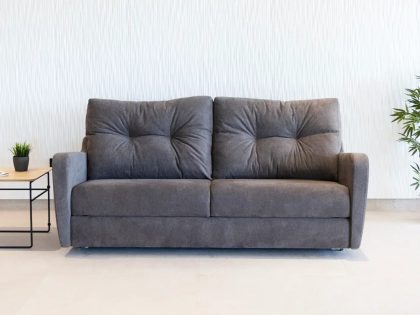 A straight-on view shows a modern dark grey two-seat sofa with plush tufted back cushions, positioned against a textured white wall with a small side table holding a plant and a taller potted plant nearby.