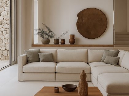 A large cream-coloured corner sofa sits in a bright, modern living room with exposed wooden ceiling beams, wooden coffee tables displaying decorative bowls and sculptures, and minimalist wall décor in warm natural tones.