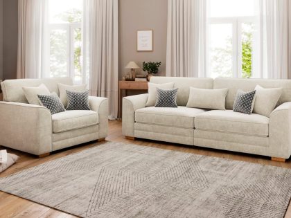 A bright, neutral-toned living room features a cream two-seater sofa and matching snuggler with patterned and plain cushions, set on a large textured rug over wooden flooring in front of tall curtained windows.