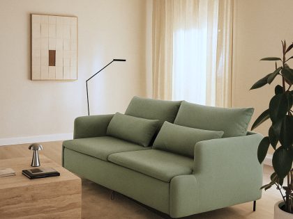 A light green two-seater sofa with matching cushions is styled in a warm, minimalist living room featuring a wooden coffee table, a floor lamp, sheer curtains, wall art, and a large potted plant.